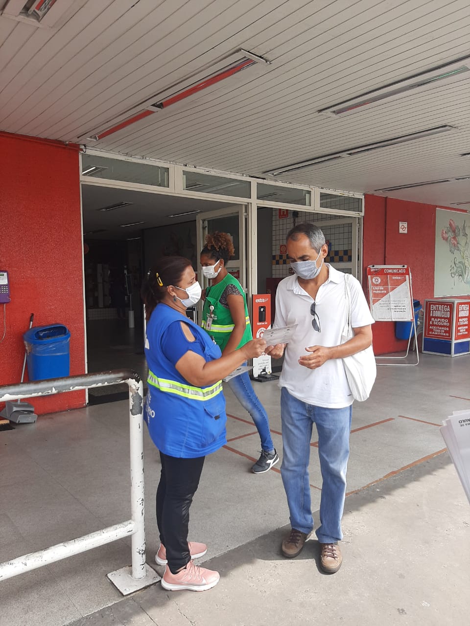 A imagem mostra uma mulher de colete azul e usando máscara interagindo com um homem de meia idade em frente a uma unidade de saúde